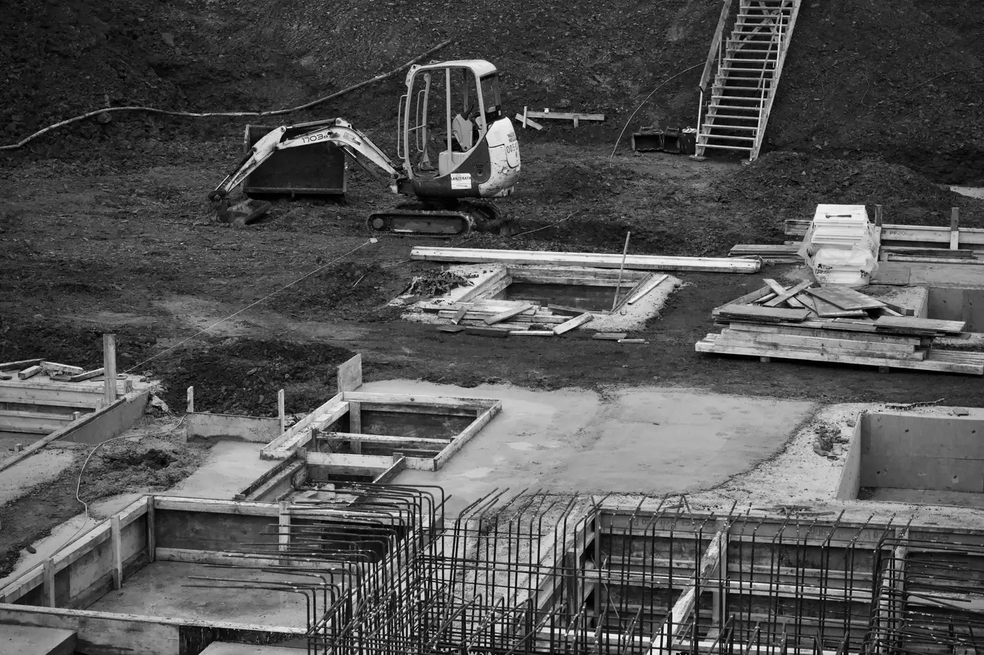 Black and white photo of an excavator at a construction site with foundation slabs and rebar laid out.