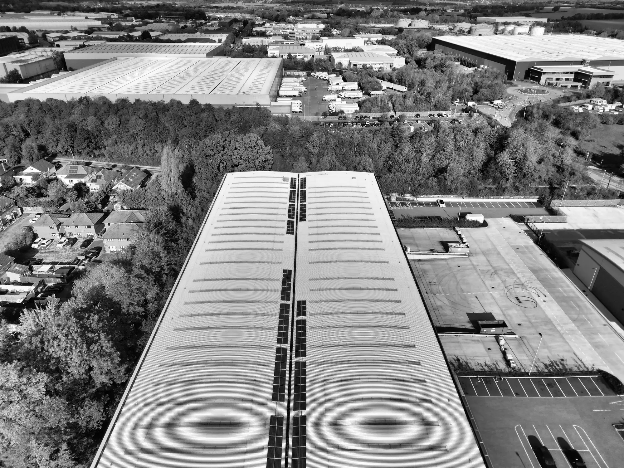 Black and white aerial view of a large single-storey building with a long ridged roof, car park and trees around it